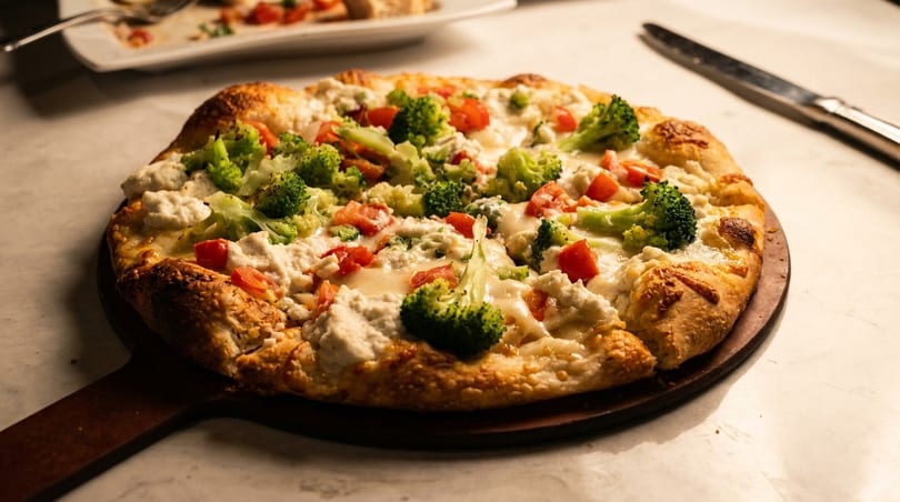 A freshly baked pizza with broccoli, tomatoes, and ricotta cheese on a wooden paddle.