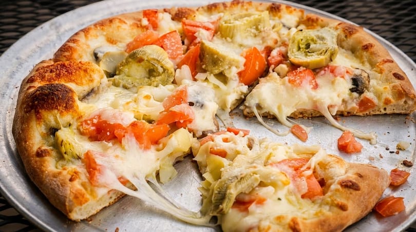A partially eaten pizza with melted cheese, tomatoes, and artichoke hearts on a metal pan.
