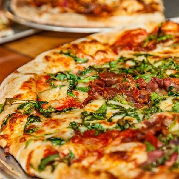 Close-up of a pizza with melted cheese, green arugula, and prosciutto on a metal tray.