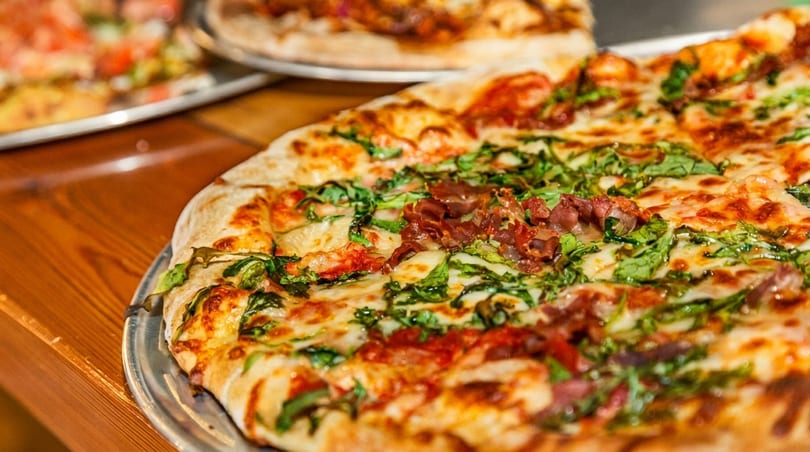 Close-up of a freshly baked pizza with melted cheese, green arugula, and prosciutto on a wooden table.