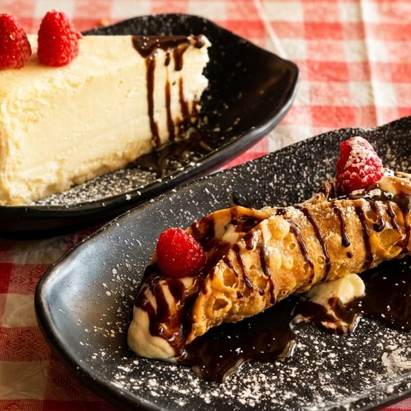 Cheesecake and a cannoli, both topped with raspberries and chocolate drizzle, on a checkered tablecloth.