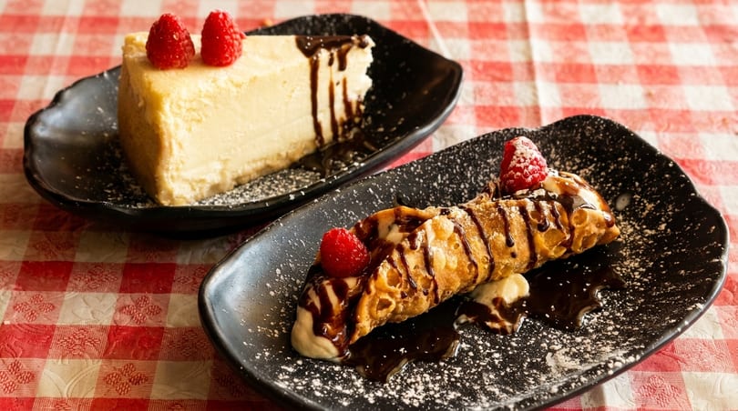 Cheesecake and cannoli with raspberries and chocolate drizzle on black plates, on a red and white checkered tablecloth.